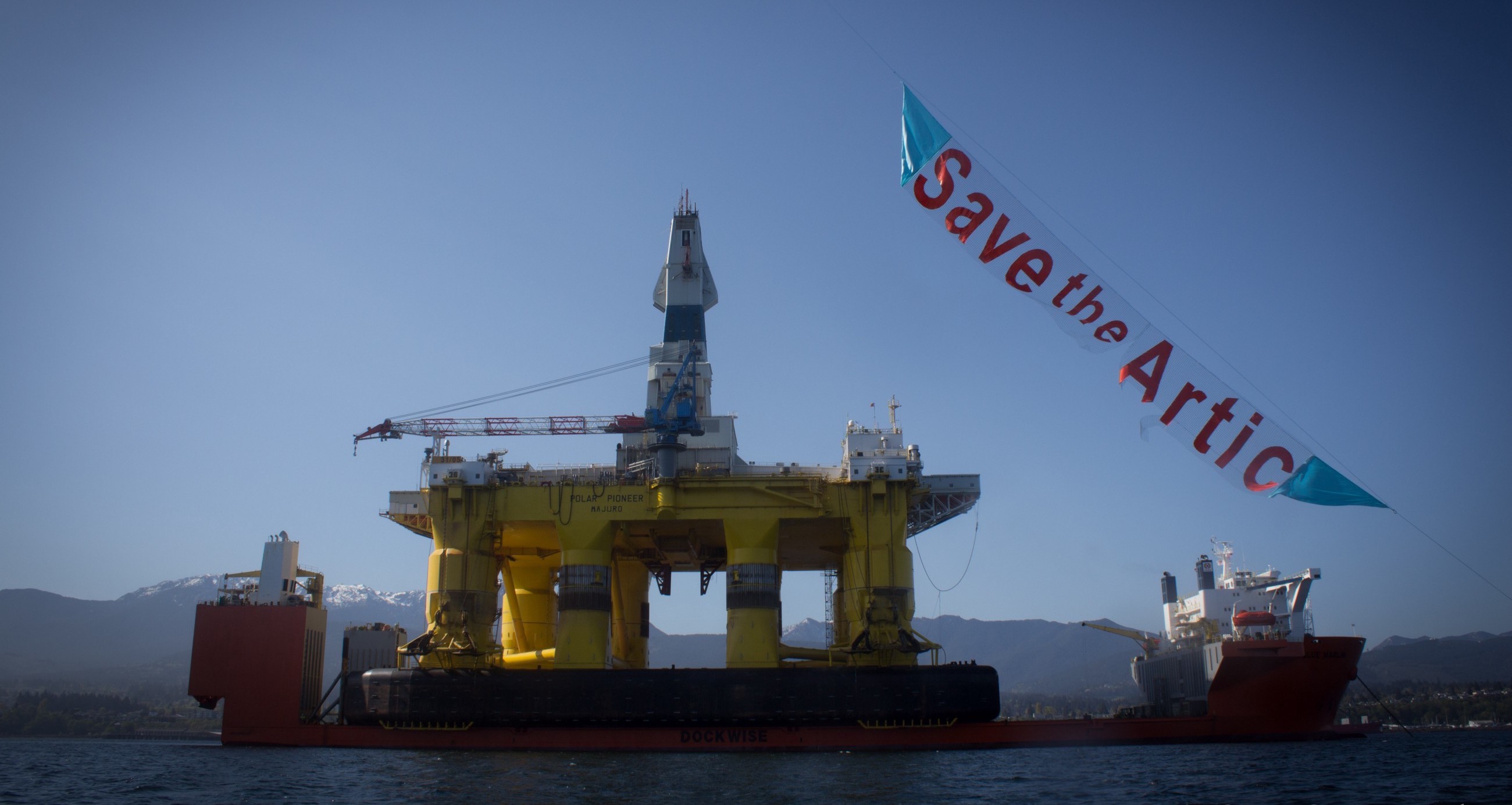 An oil platform sits on a ship, awaiting its voyage out to sea. Activists fly a "Save the Arctic" banner overhead.