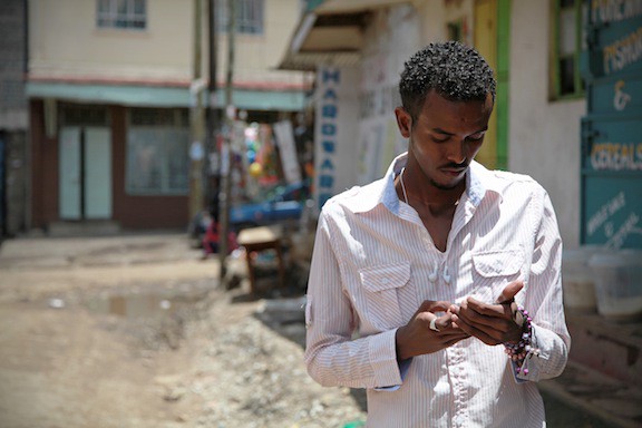 A man stands looking into his smartphone in a dustry unpaved street.