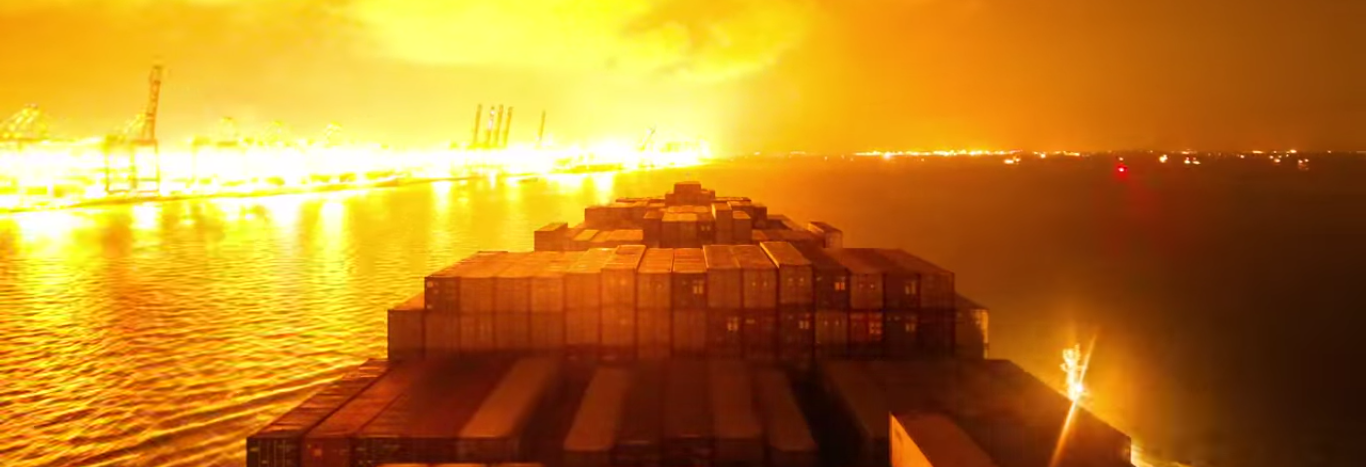 A container ship sailing at night along a shoreline lit up with extensive, bright artificial lights from docks and cities