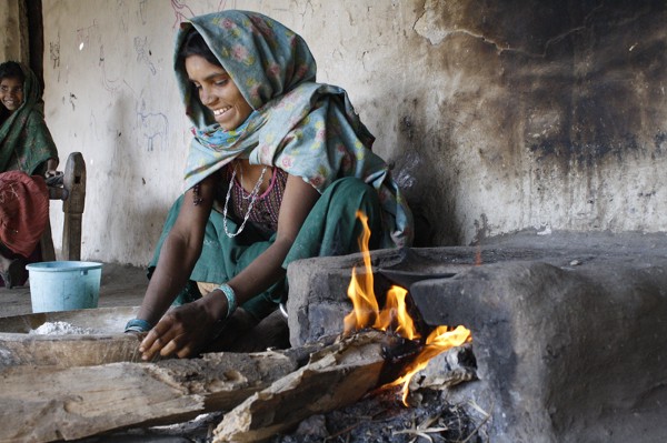 A woman in a shawl is picking up firewood to place into the stove next to her.